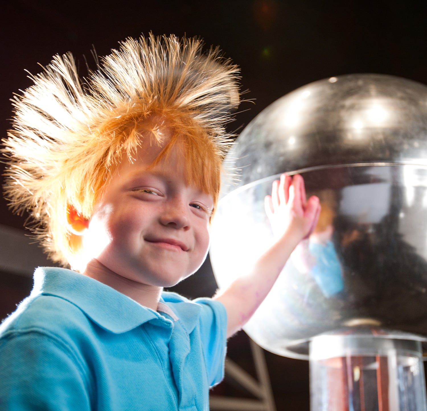 Child explores electricity as hair stands up touching Van de Graaff generator at children's science center.