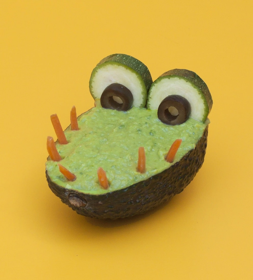 Kids food science craft: avocado crocodile with guacamole, carrot teeth, and olive eyes on yellow background