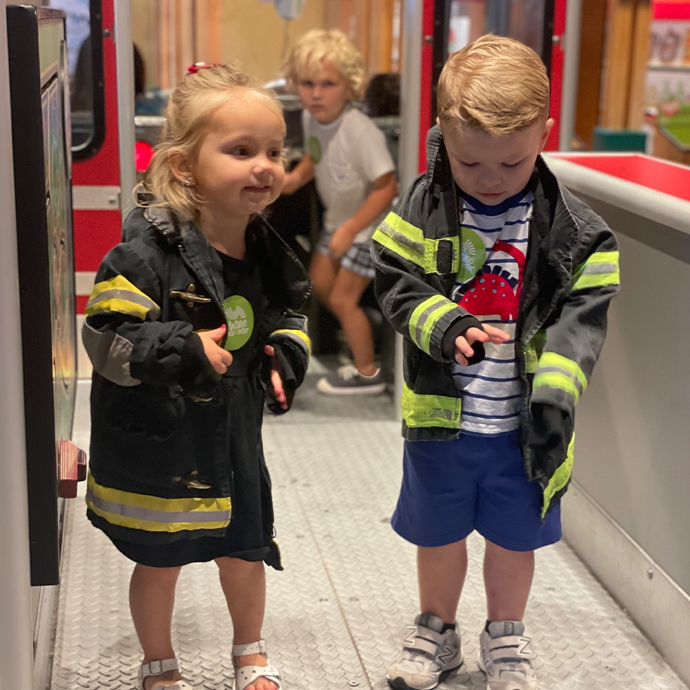 Kids in firefighter jackets exploring a fire truck exhibit at a children's science center, engaging in pretend play.