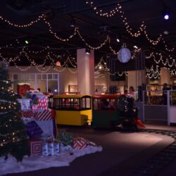 Holiday train ride at children's science center with lights, Christmas tree, gifts, and a child riding the mini locomotive.