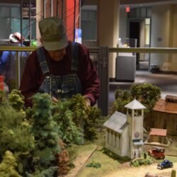A person in engineer cap tending a model train village at children's science center exhibit with mini church and trees