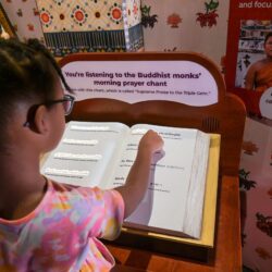 Child explores interactive exhibit, listening to Buddhist monks’ morning chant and reading a large book at a science center.