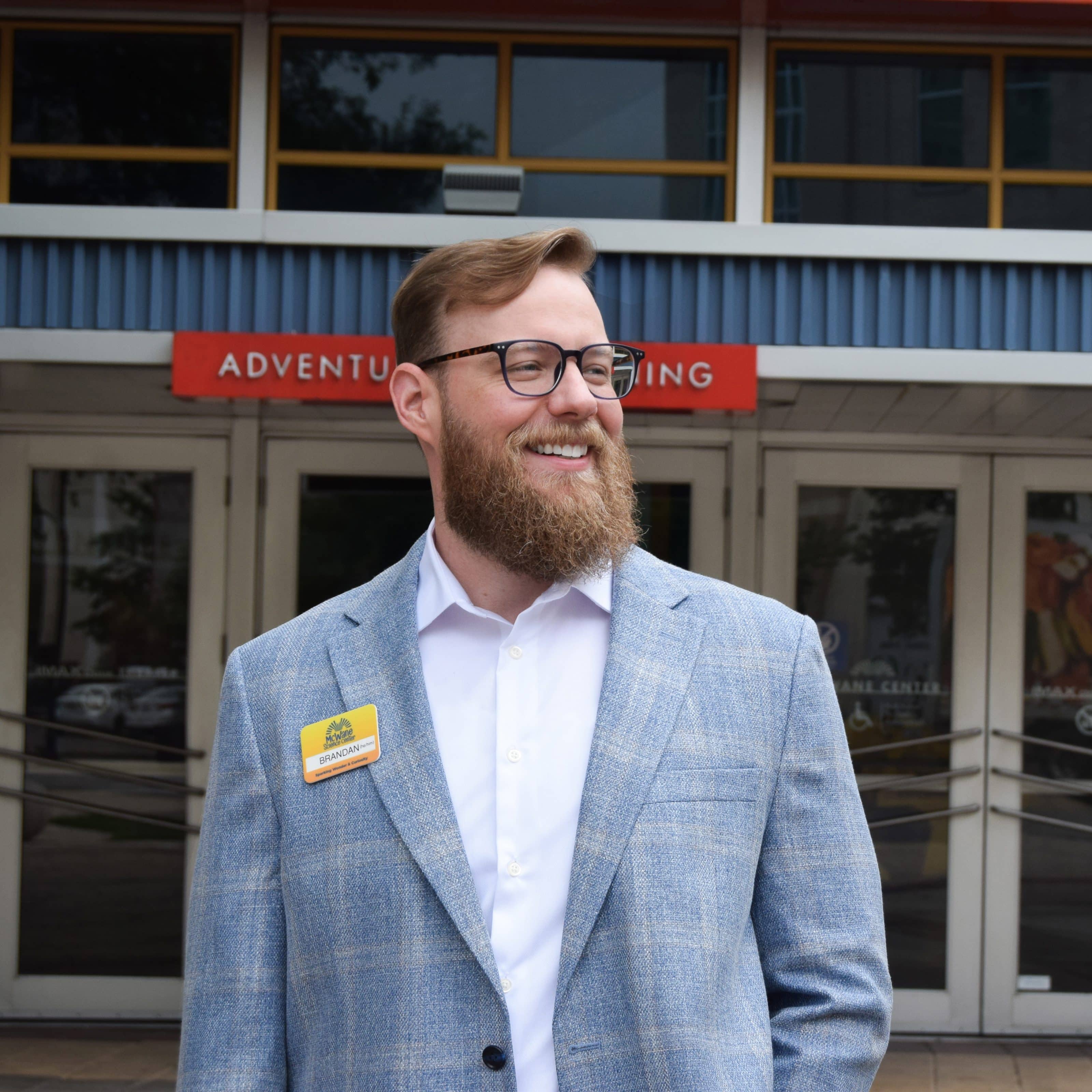 Smiling staff member at children's science center entrance, wearing blazer and name badge, welcoming families.