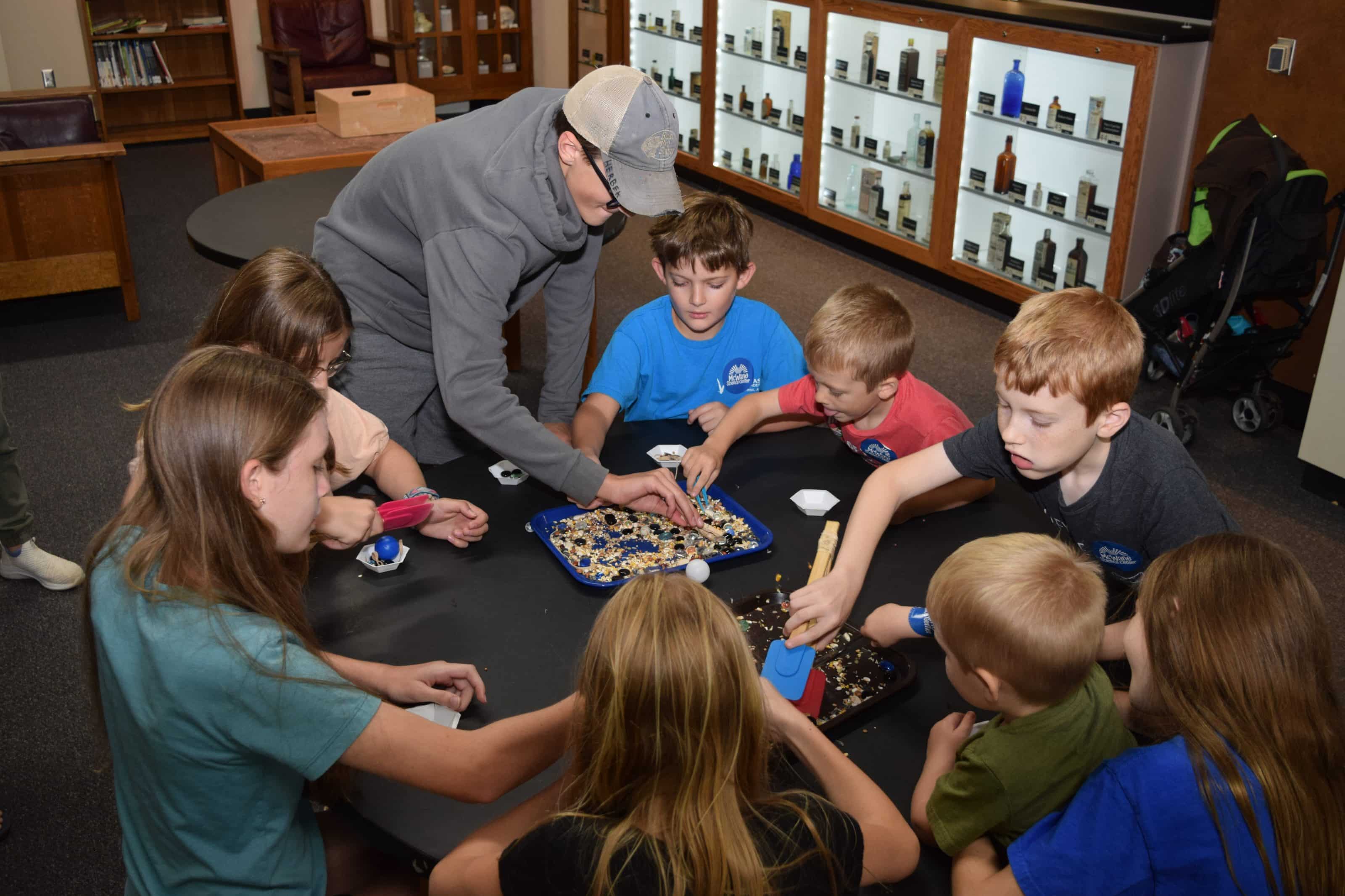 Children and an educator explore a hands-on STEM activity at a science center, sorting materials with scoops and tools.