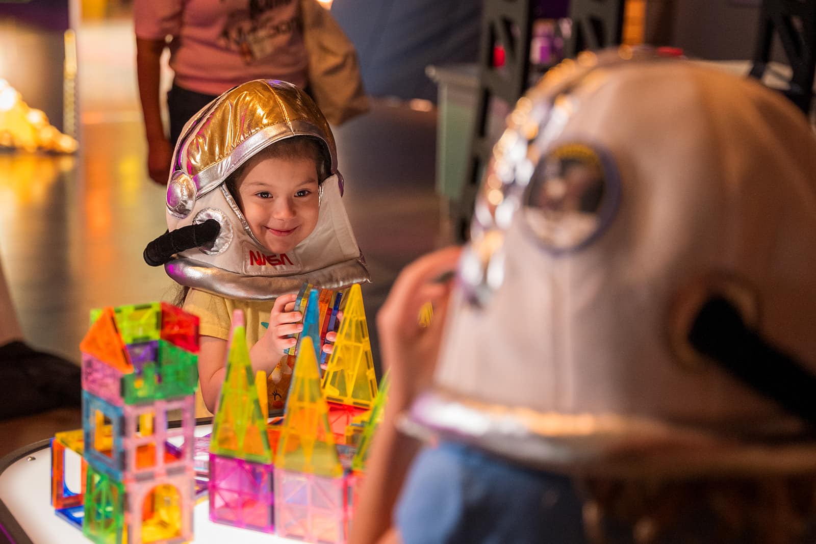 Kids in astronaut helmets build colorful magnetic tiles at a children's science center STEM exhibit