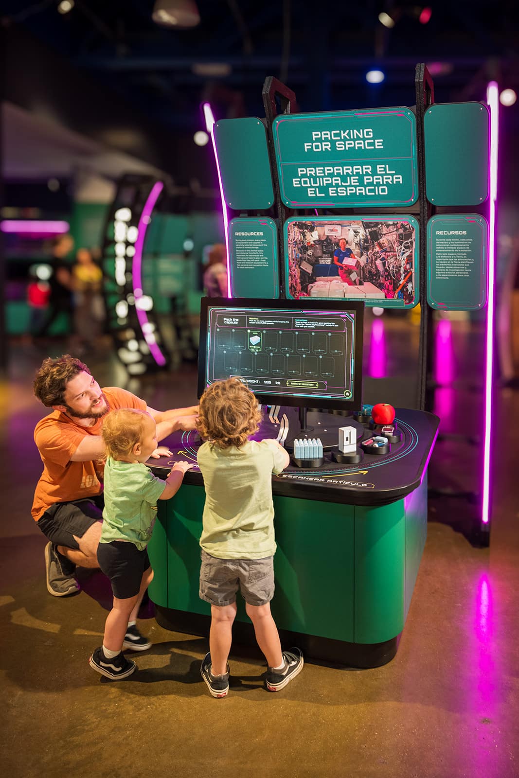 Kids explore Packing for Space interactive exhibit at a children's science center with an adult helper.