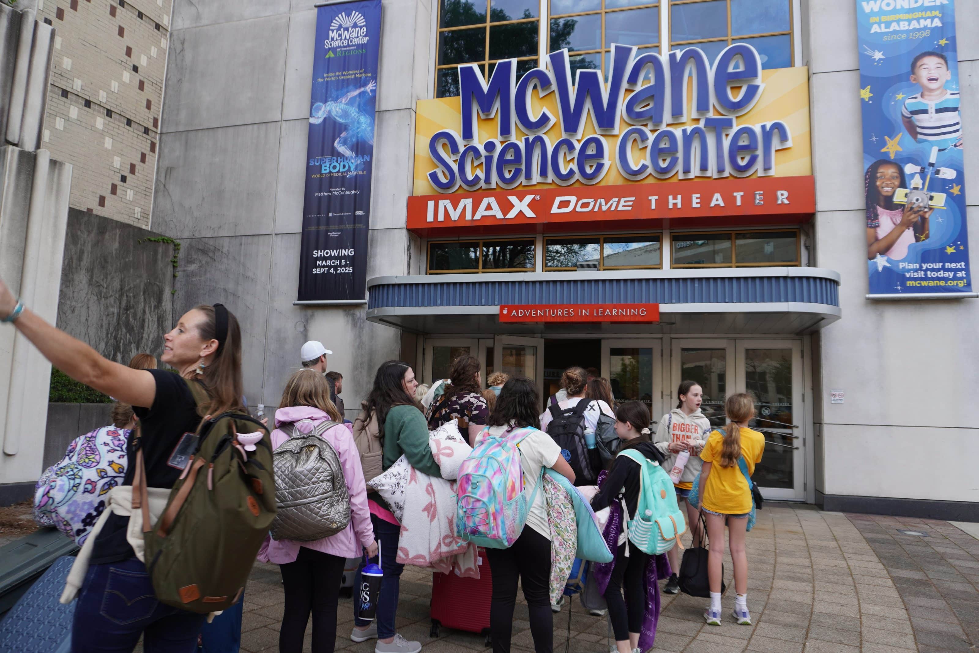 Kids and a teacher enter McWane Science Center IMAX Dome Theater for a field trip, backpacks and pillows at the entrance.