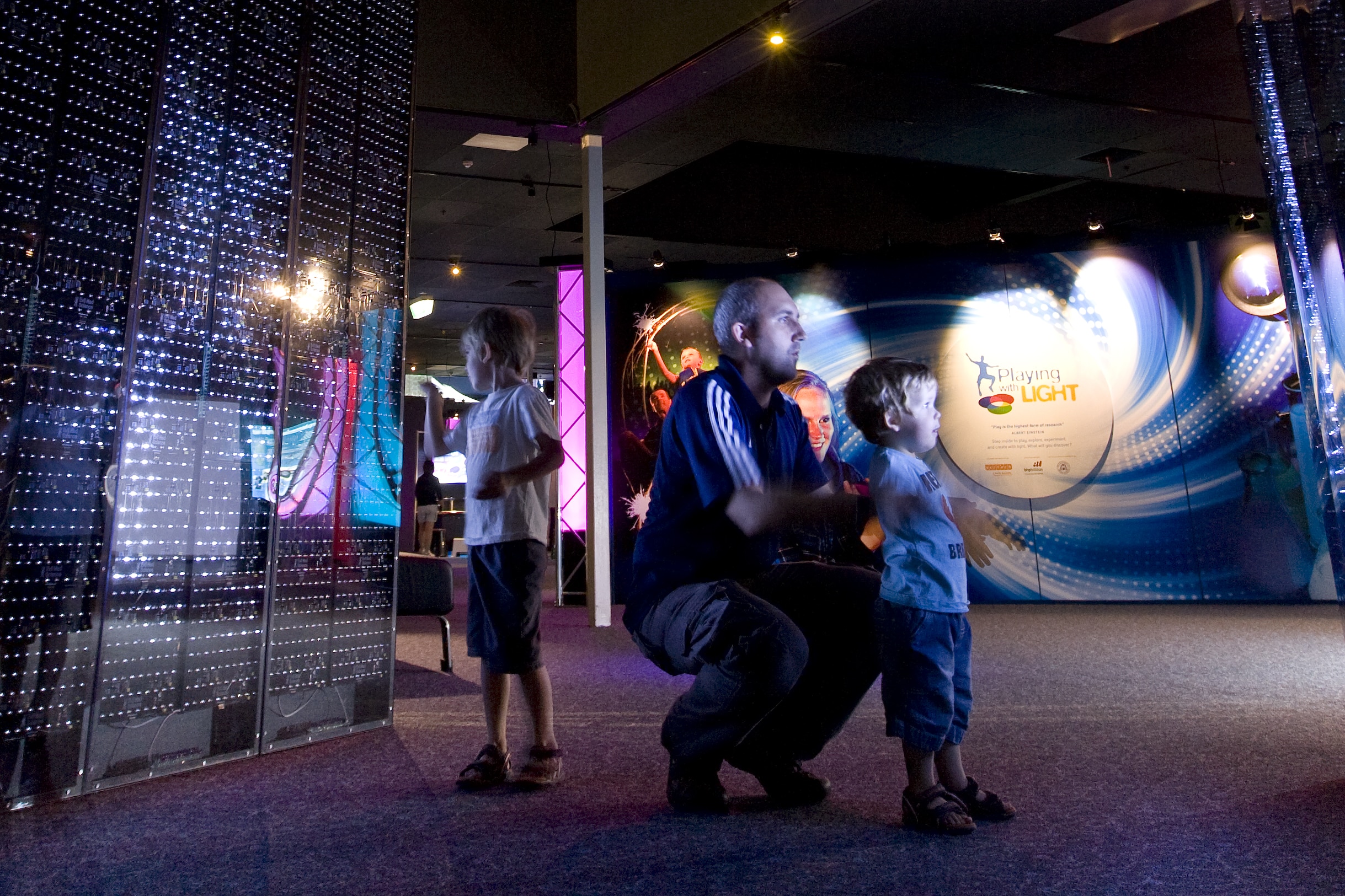 A man and children explore the Playing with Light interactive exhibit at a children's science center.