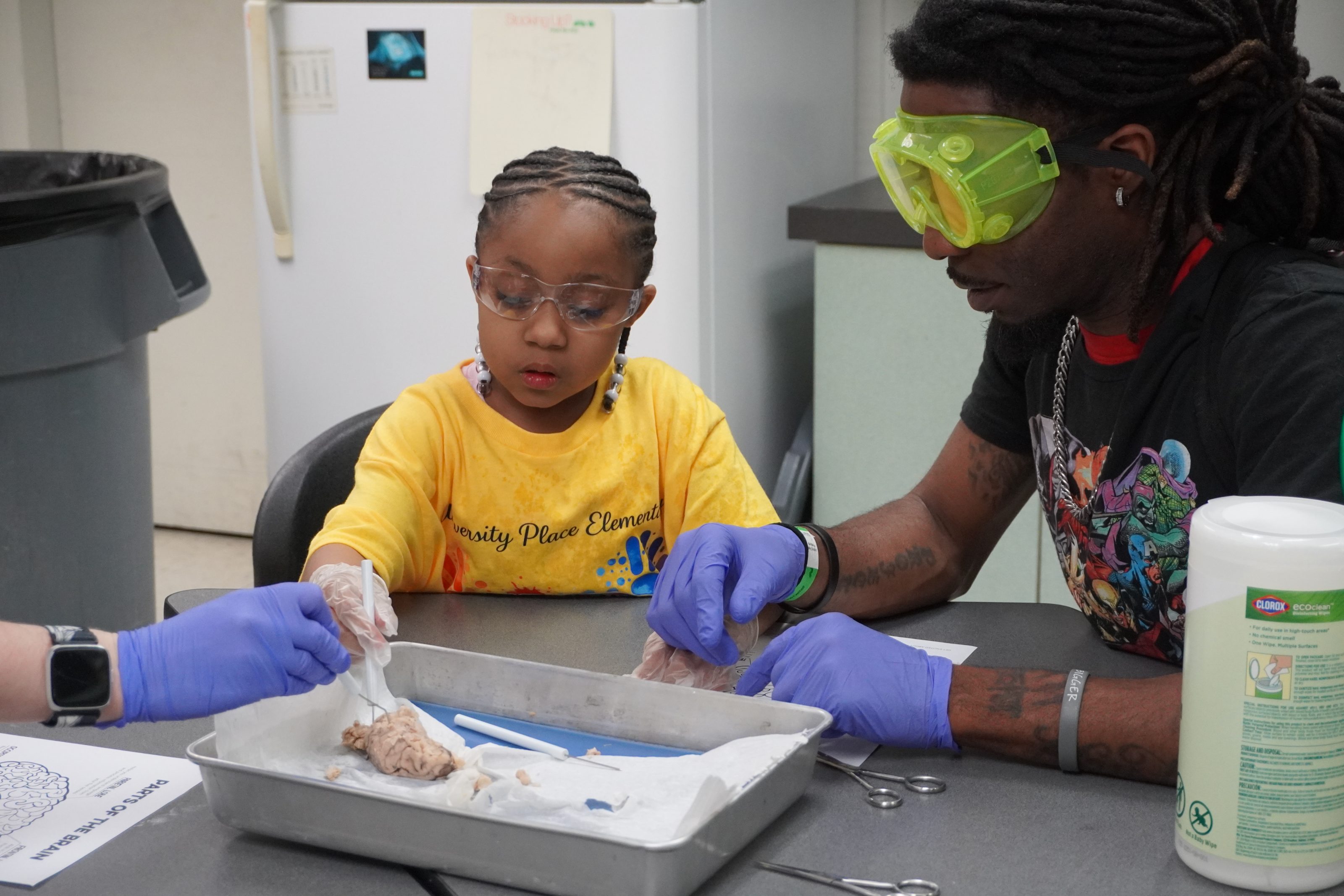 A young student wearing protective gloves and goggles examines a brain specimen with help from an educator.